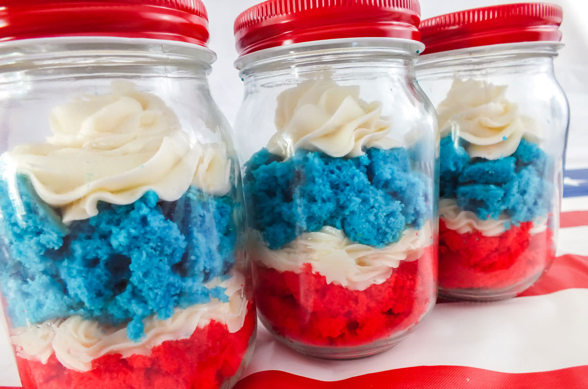 Three Red White and Blue Cupcakes in Jar sitting on a red striped table cloth in front of a white background.