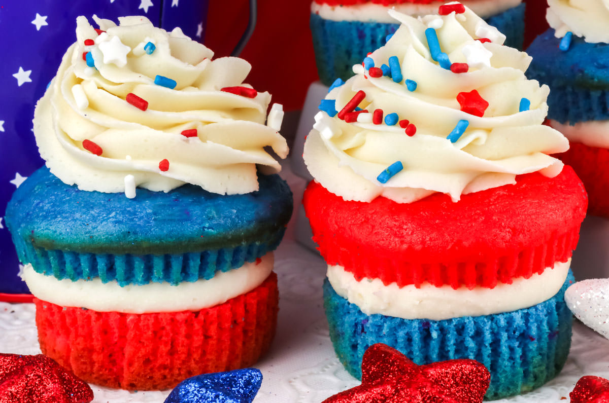 Close up on two Red White and Blue Cupcakes sitting on a white surface in front of 4th of July decorations.