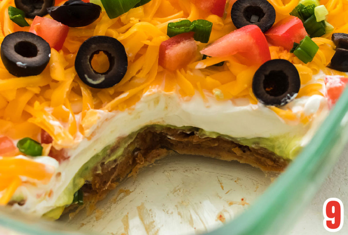 Closeup on a clear glass dish of 7 Layer Dip with a section scooped away so you can see all seven layers of the dip.