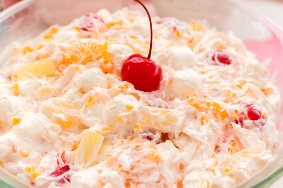 Closeup on a glass serving bowl filled with Ambrosia Salad and topped with a Maraschino Cherry with a stem on it.