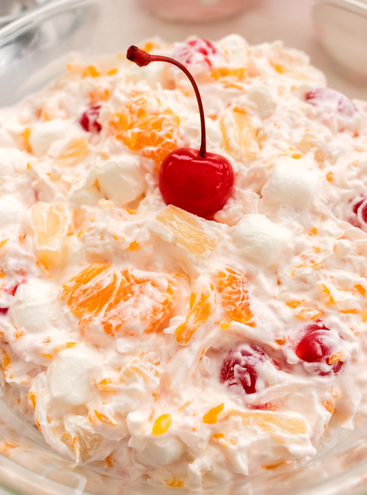 Closeup on a glass bowl filled with Ambrosia Salad and topped with a Maraschino Cherry.