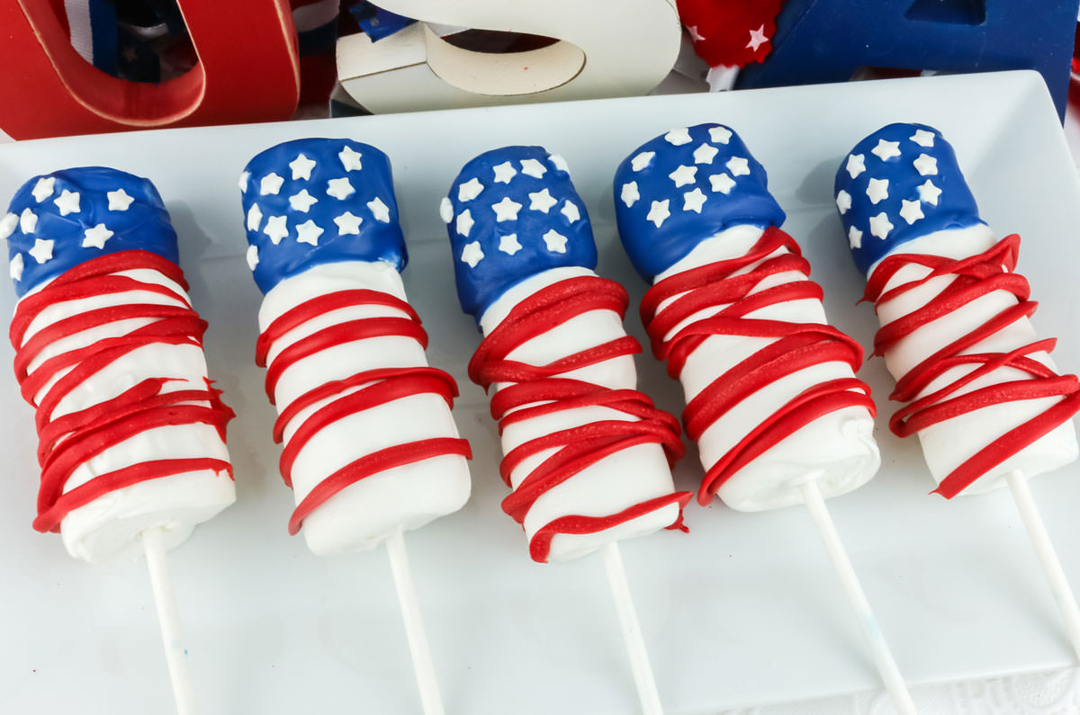 Five American Flag Marshmallow Pops laying on a white serving platter in front of at red white and blue USA sign.