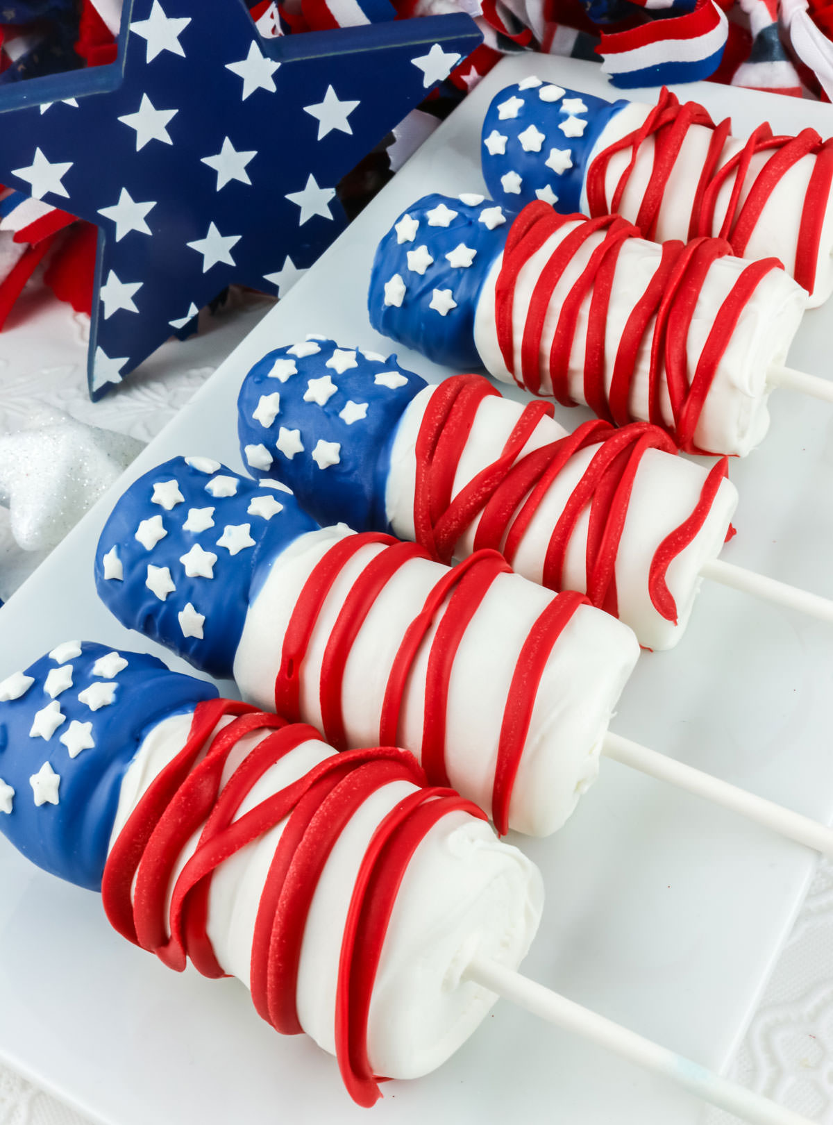 Five red white and blue American Flag Marshmallow Pops laying on a white serving platter on a white table in front of 4th of July decorations.