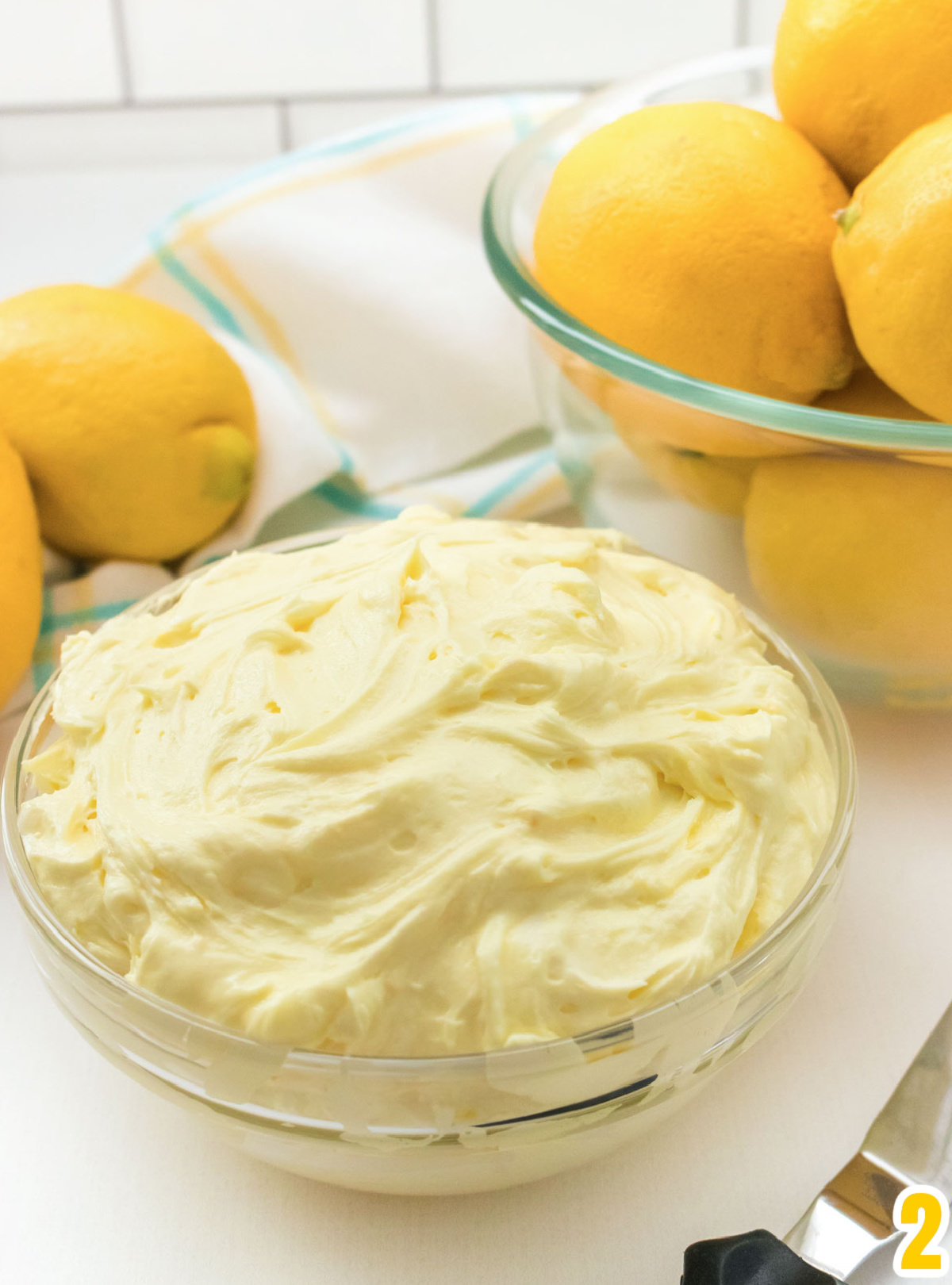 A glass bowl filled with Lemon Frosting sitting in front of a glass bowl filled with Lemons.
