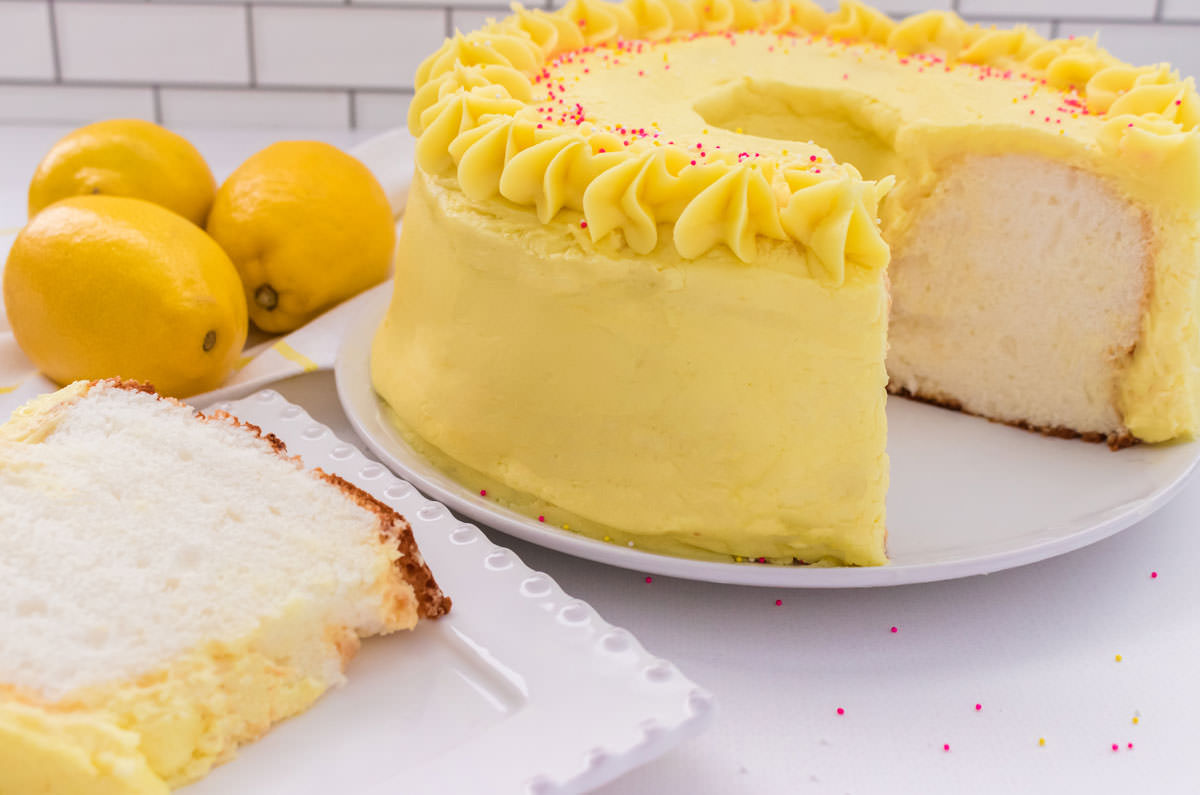 Closeup of an Angel Food Cake with Lemon Frosting on a cake plate on a white table with a slice taken out and on a dessert plate.