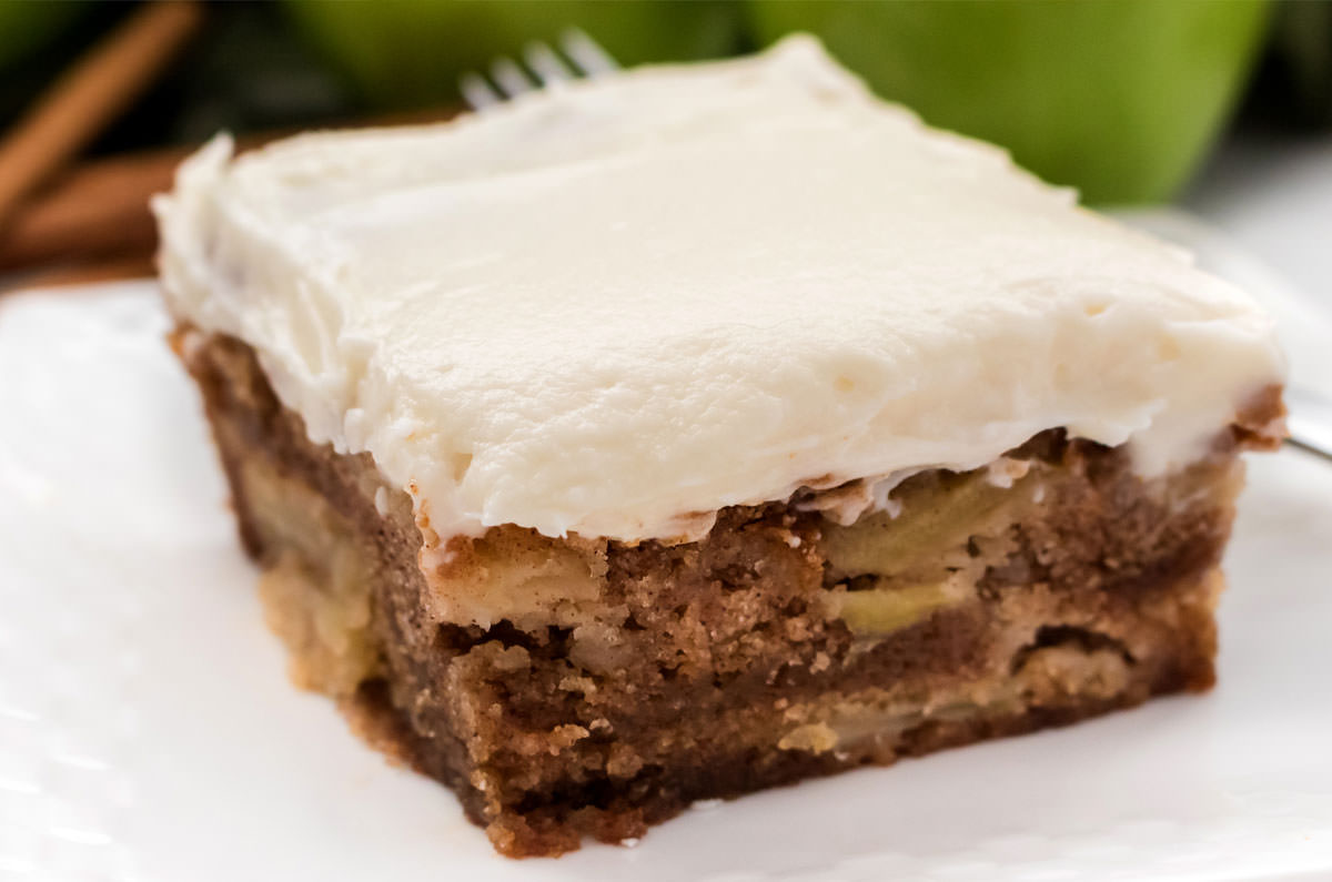 Closeup on a single slice of Apple Cake sitting on a white plate in front of a stack of apples.