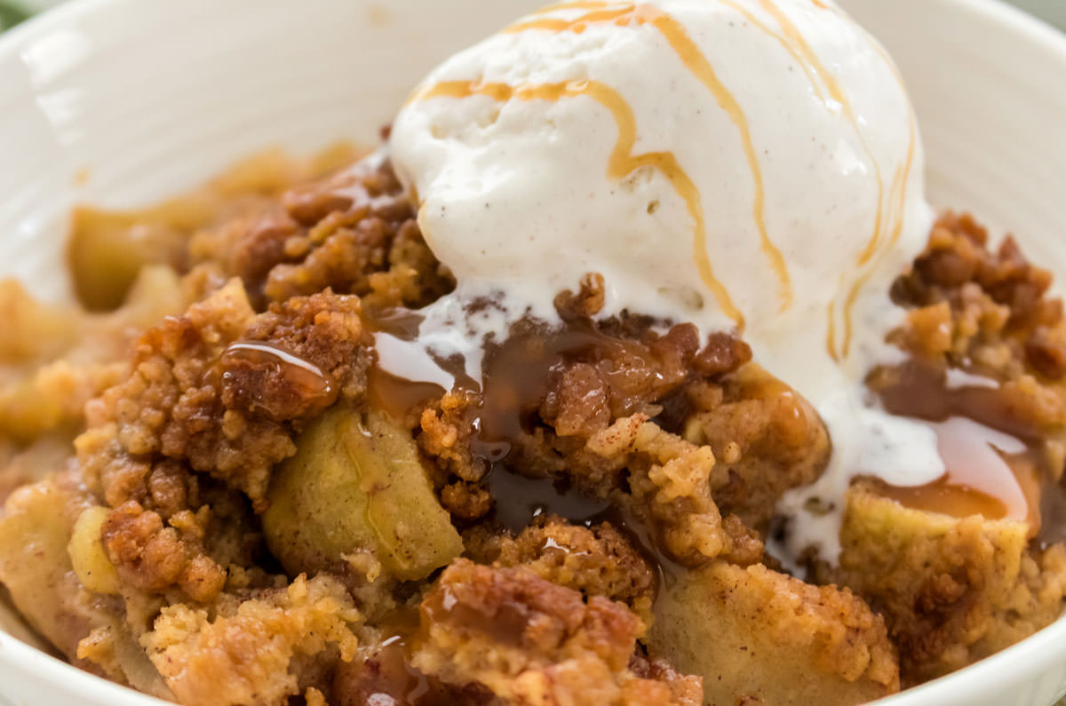 Close on a white bowl of Easy Apple Cobbler straight out of the oven topped with ice cream and caramel sauce.
