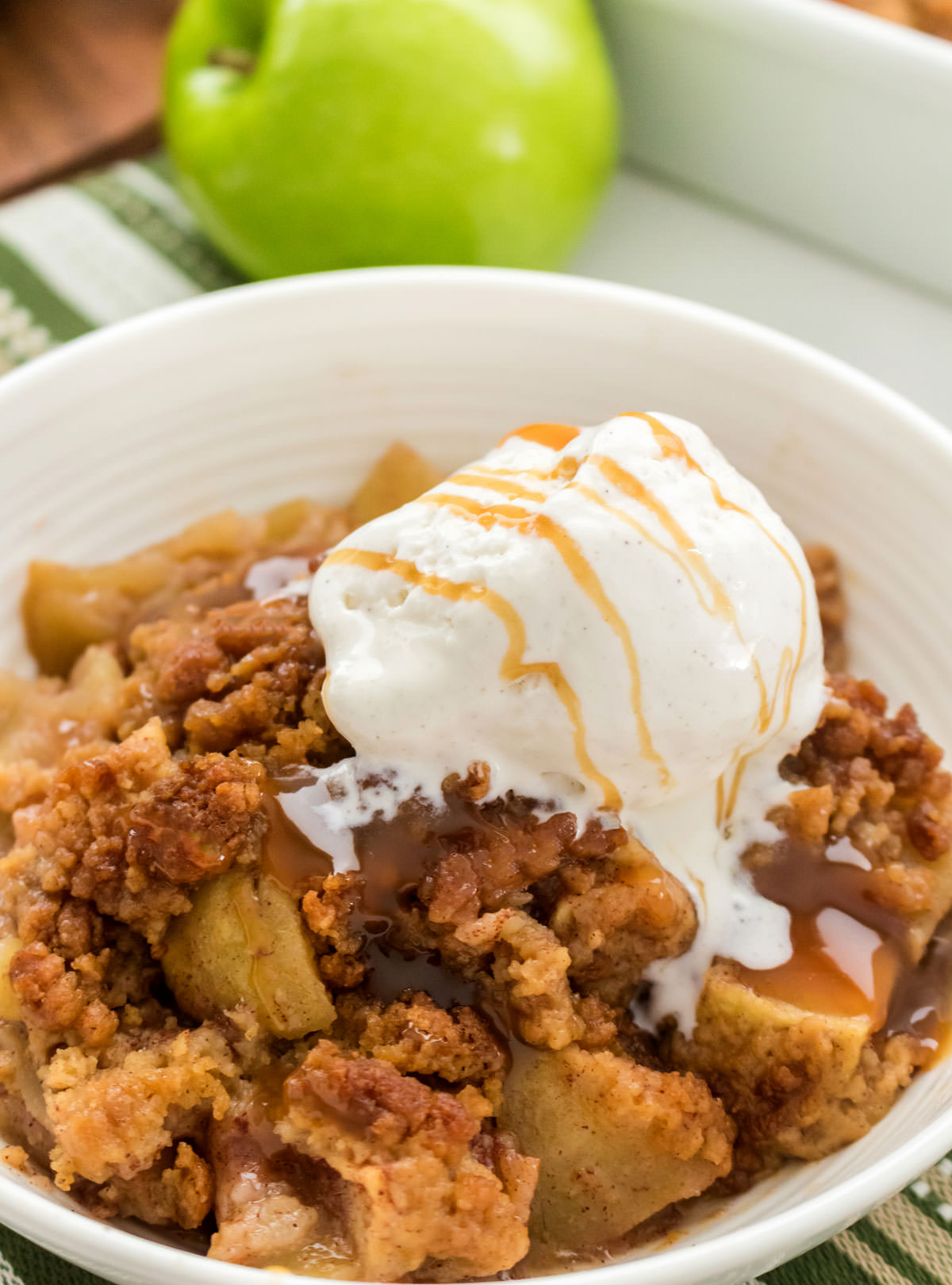 Closeup on a white bowl filled with homemade Apple Cobbler with a scoop of Vanilla Ice Cream that has been drizzled with caramel syrup.