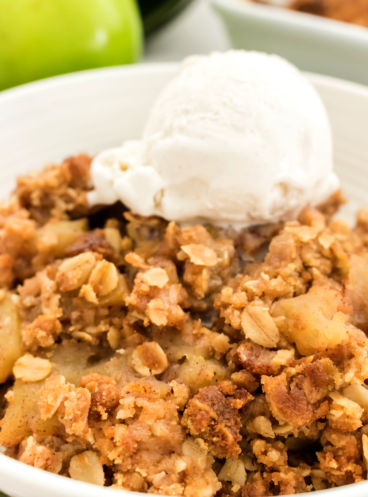 Closeup on a white bowl filled with warm Apple Crisp topped with a scoop of Vanilla Ice Cream.