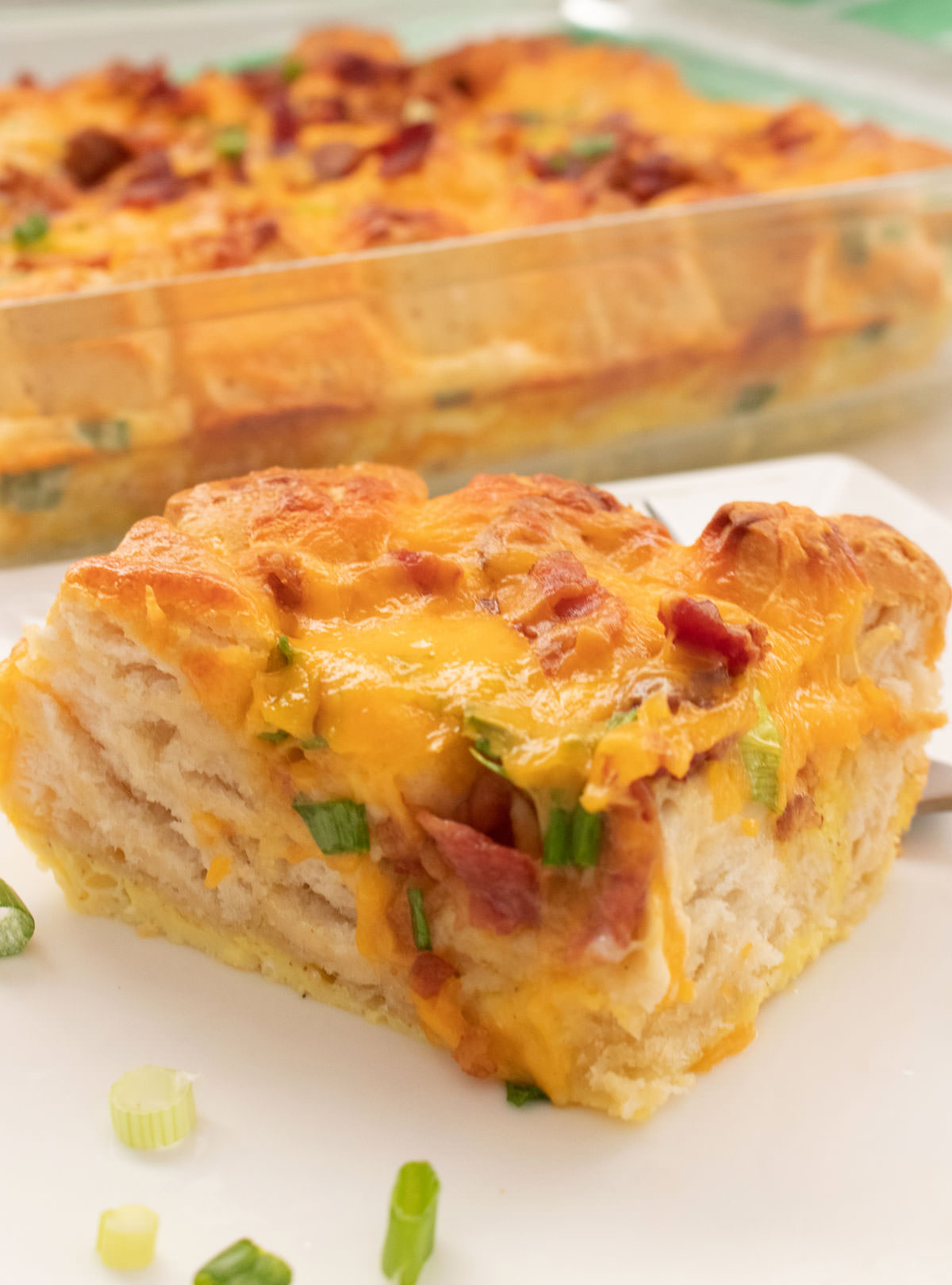 A close up of a piece of Bacon and Egg Breakfast Casserole sitting on a white plate in front of the casserole pan.