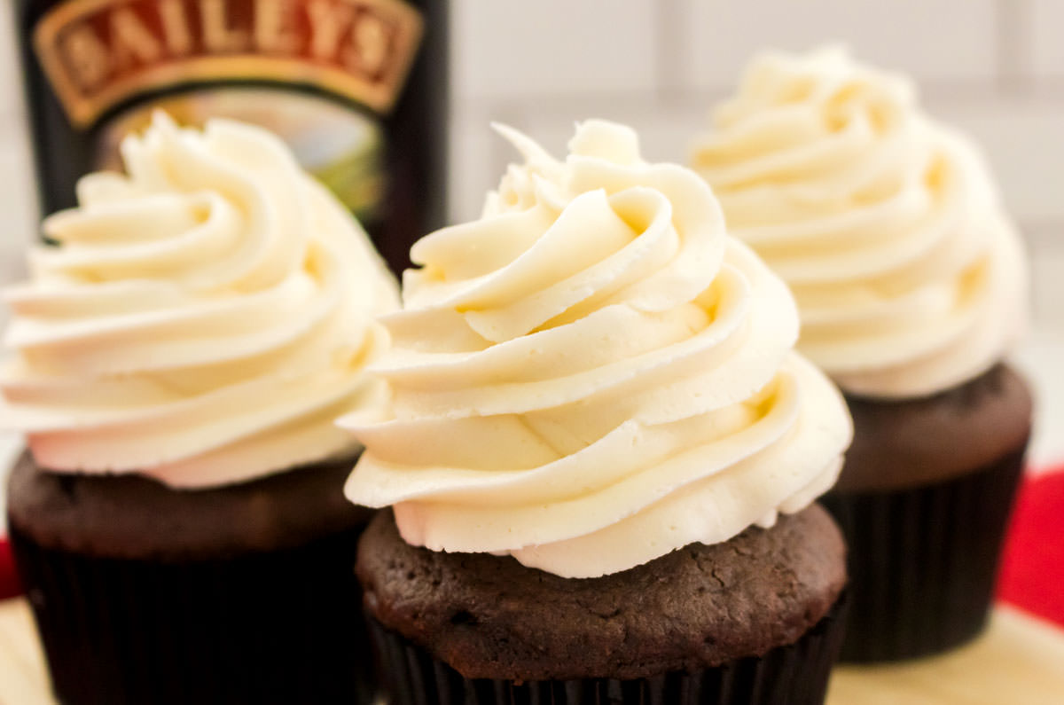 Closeup on three cupcakes topped with Baileys Irish Cream Buttercream Frosting sitting on a cutting board in front of a bottle of the liqueur. 