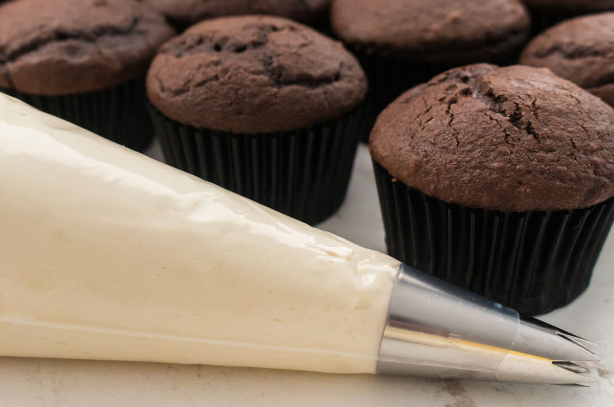 Closeup on a decorating bag filled with Baileys Irish Cream Frosting sitting in front of a batch of unfrosted chocolate cupcakes.