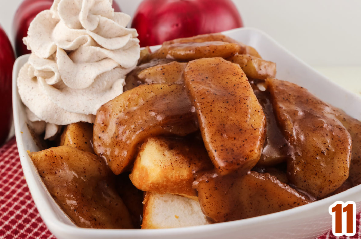 Closeup on a white serving bowl sitting on a red kitchen towel filled with Cinnamon Apples topped with Whipped Cream.