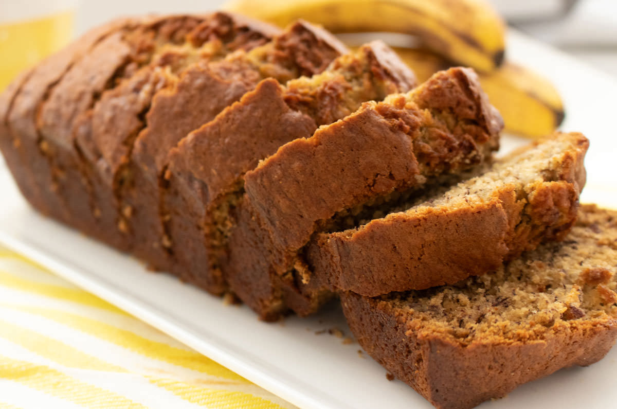 Closeup of a loaf of Banana Bread laying on a white serving platter on a yellow tea towel in front of a bunch of ripe bananas.