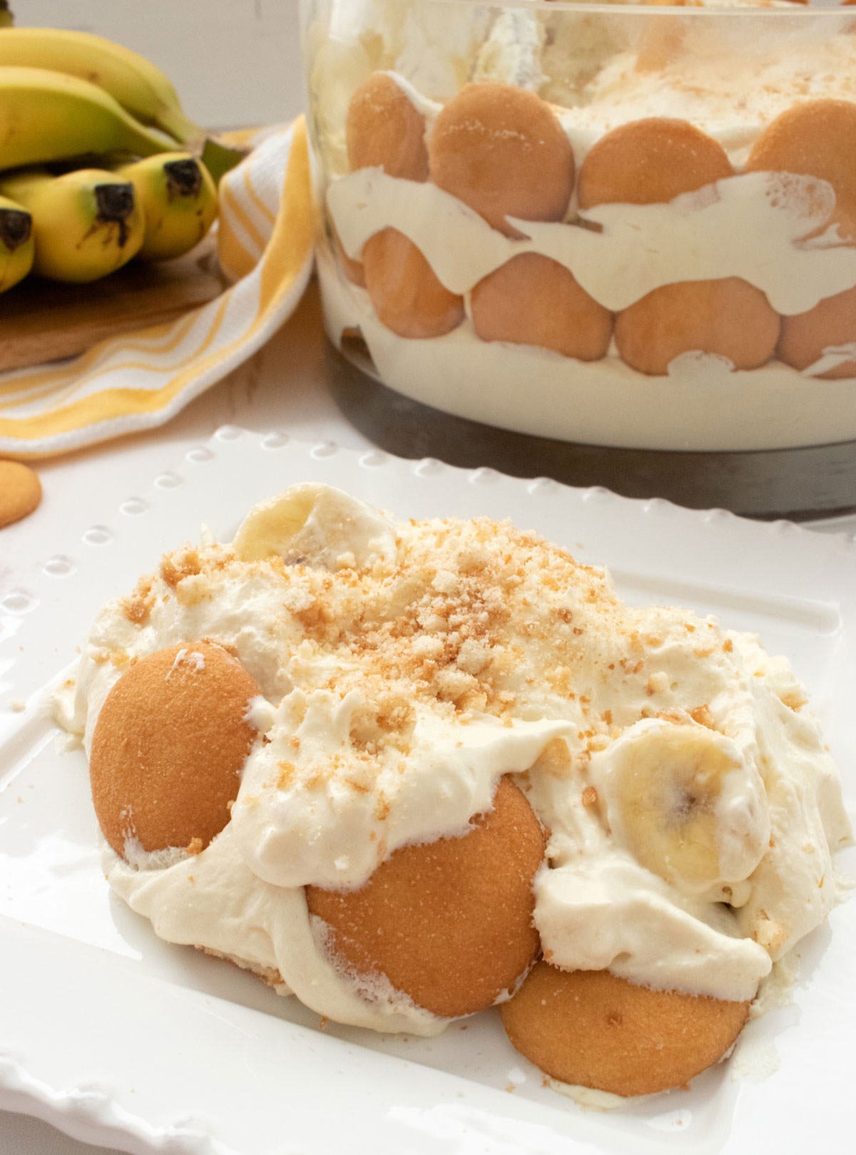 A serving of Banana Pudding on a white plate in front of the big bowl of the Banana Pudding. 
