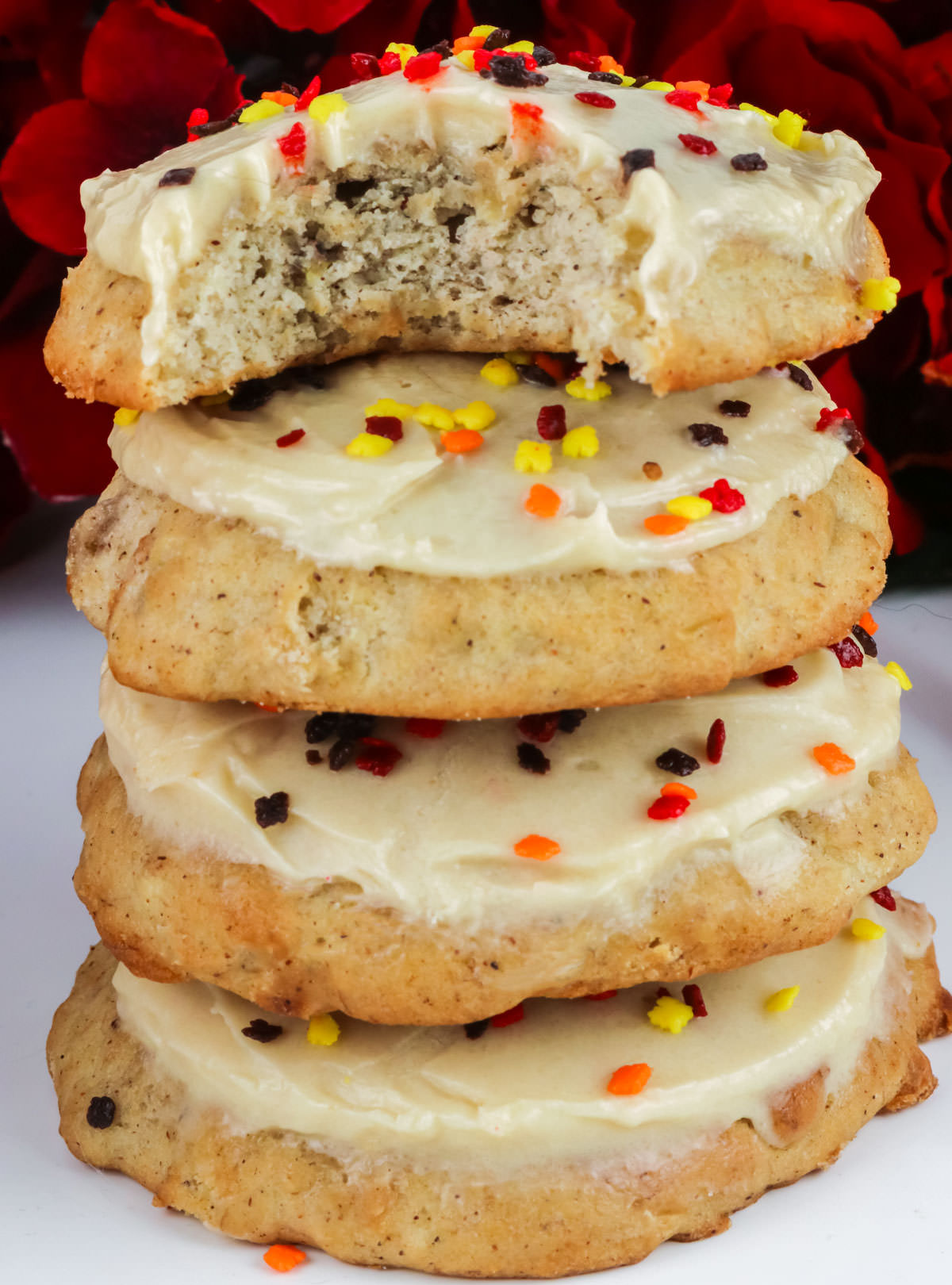 Closeup on a stack of Banana Spice Cookies with Brown Sugar Cream Cheese Frosting sitting on a white table in front of red flowers.