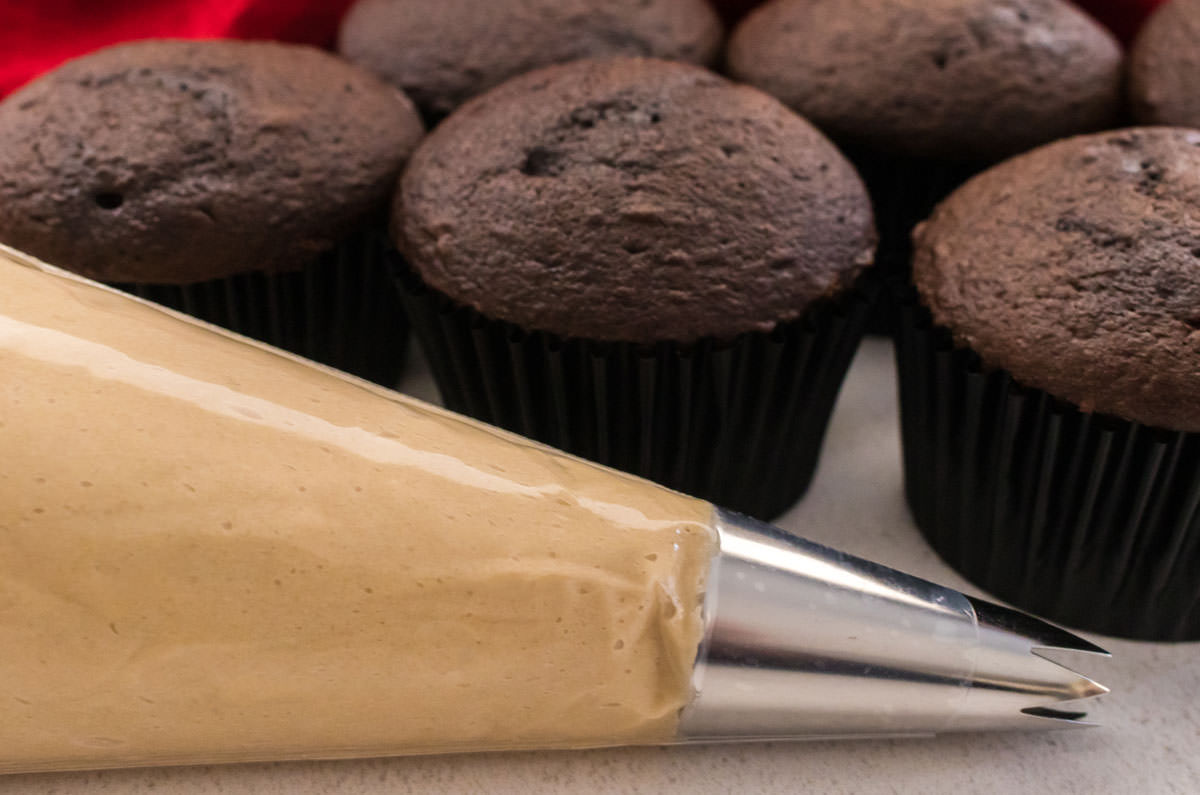 Closeup on a decorating bag filled with Caramel Frosting sitting in front of a batch of unfrosted Chocolate Cupcakes.