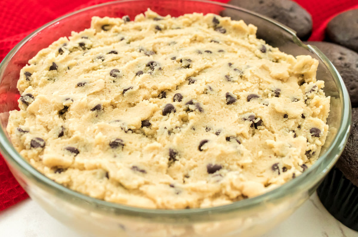 A glass bowl filled with Cookie Dough Frosting sitting next to a batch of unfrosted chocolate cupcakes.