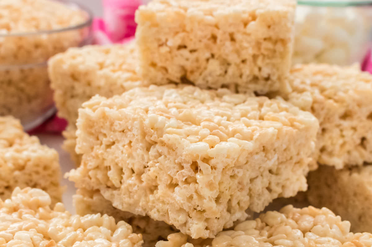 Closeup on a stack of The Best Ever Rice Krispie Treats sitting on a white table in front of a bowl of marshmallows and a bowl of Rice Krispie Cereal.