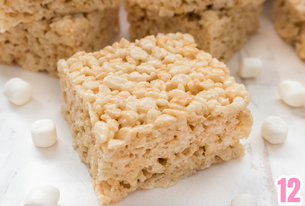 Closeup of a single Rice Crispy Treat sitting on a white surface surrounded by mini marshmallows.