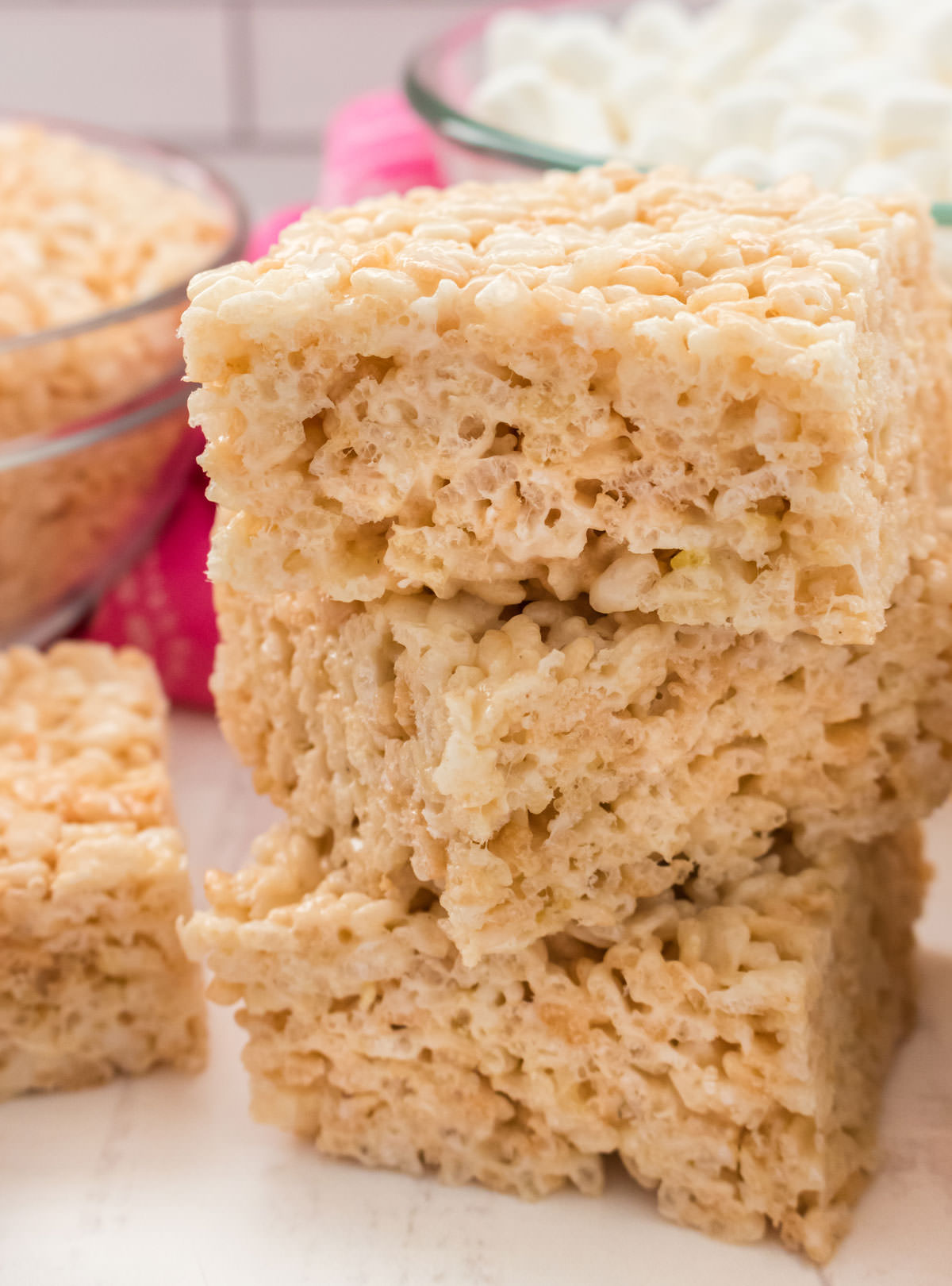 Closeup on a stack of three Rice Krispie Treats sitting on a white table next to a clear glass bowl filled with Rice Krispie Cereal.