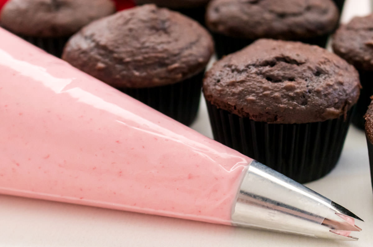 Closeup on a decorating bag filled with Raspberry Frosting sitting in front of a batch of unfrosted chocolate cupcakes.