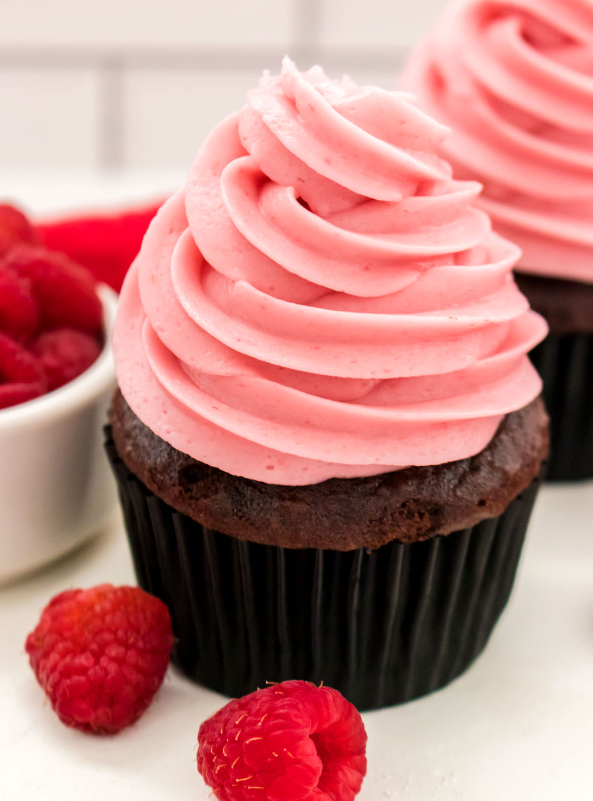 Closeup on a cupcake topped with The Best Raspberry Buttercream Frosting sitting next to a ramekin filled with Fresh Raspberries.