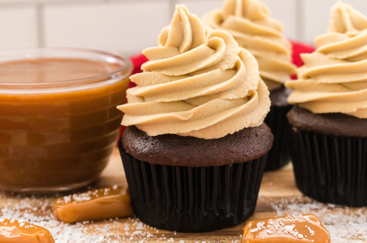 Closeup on three chocolate cupcakes topped with The Best Salted Caramel Buttercream Frosting sitting next to a glass bowl filled with caramel sauce.