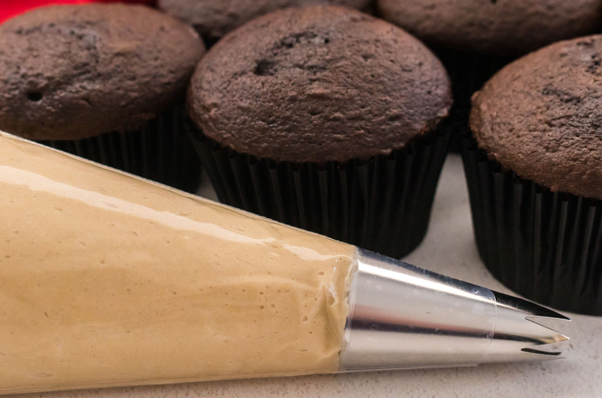 Closeup on a decorating bag filled with Salted Caramel Frosting sitting on a white table in front of a batch of chocolate cupcakes.