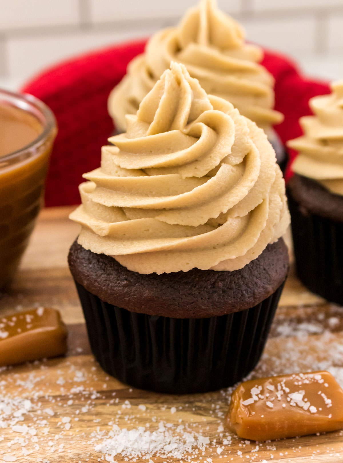 Closeup on three chocolate cupcakes topped with The Best Salted Caramel Buttercream Frosting sitting on a cutting board surrounded by Sea Salt and Caramel candies.