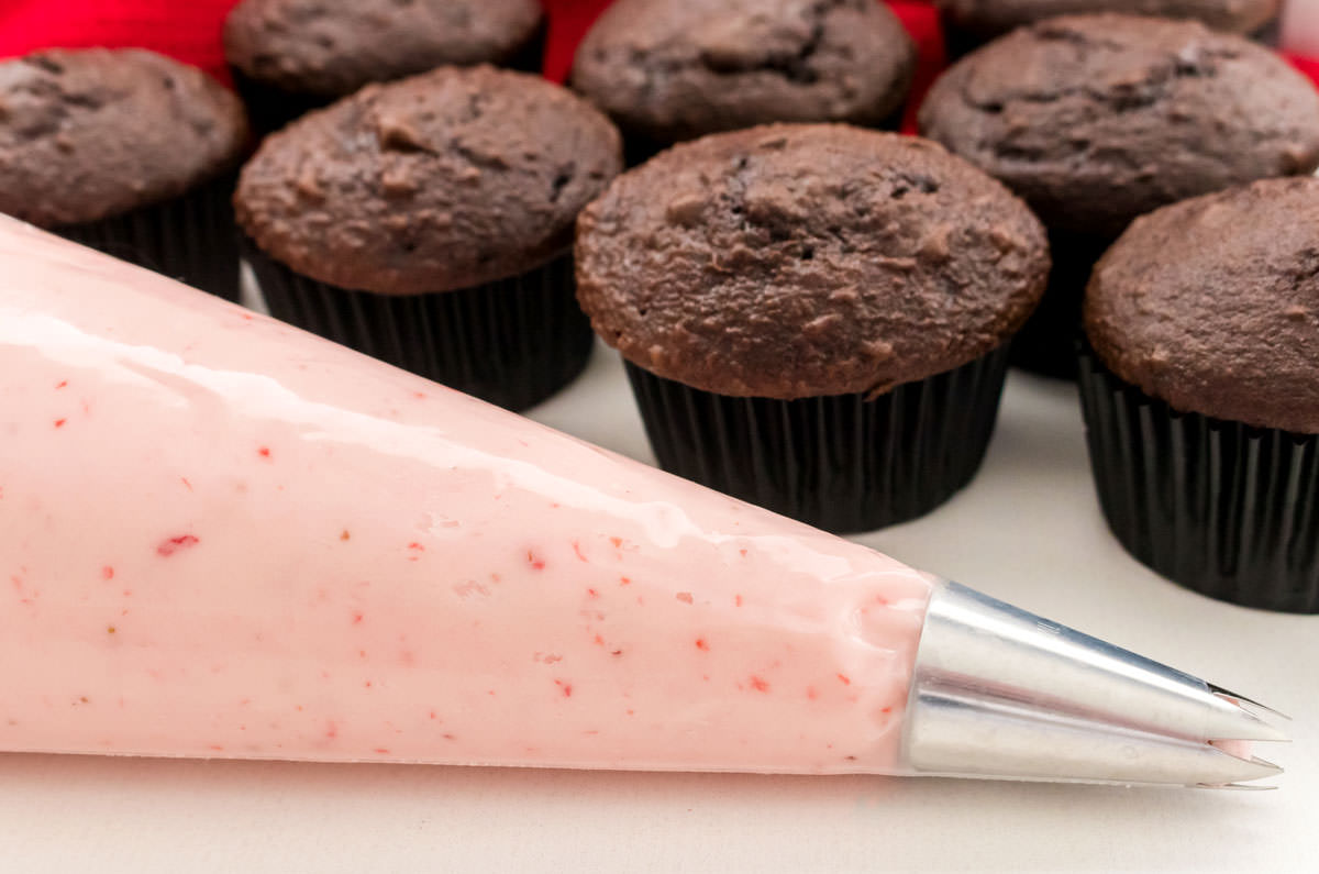 Closeup on a decorating bag filled with homemade Strawberry Frosting sitting on a white table in front of a batch of unfrosted chocolate cupcakes.