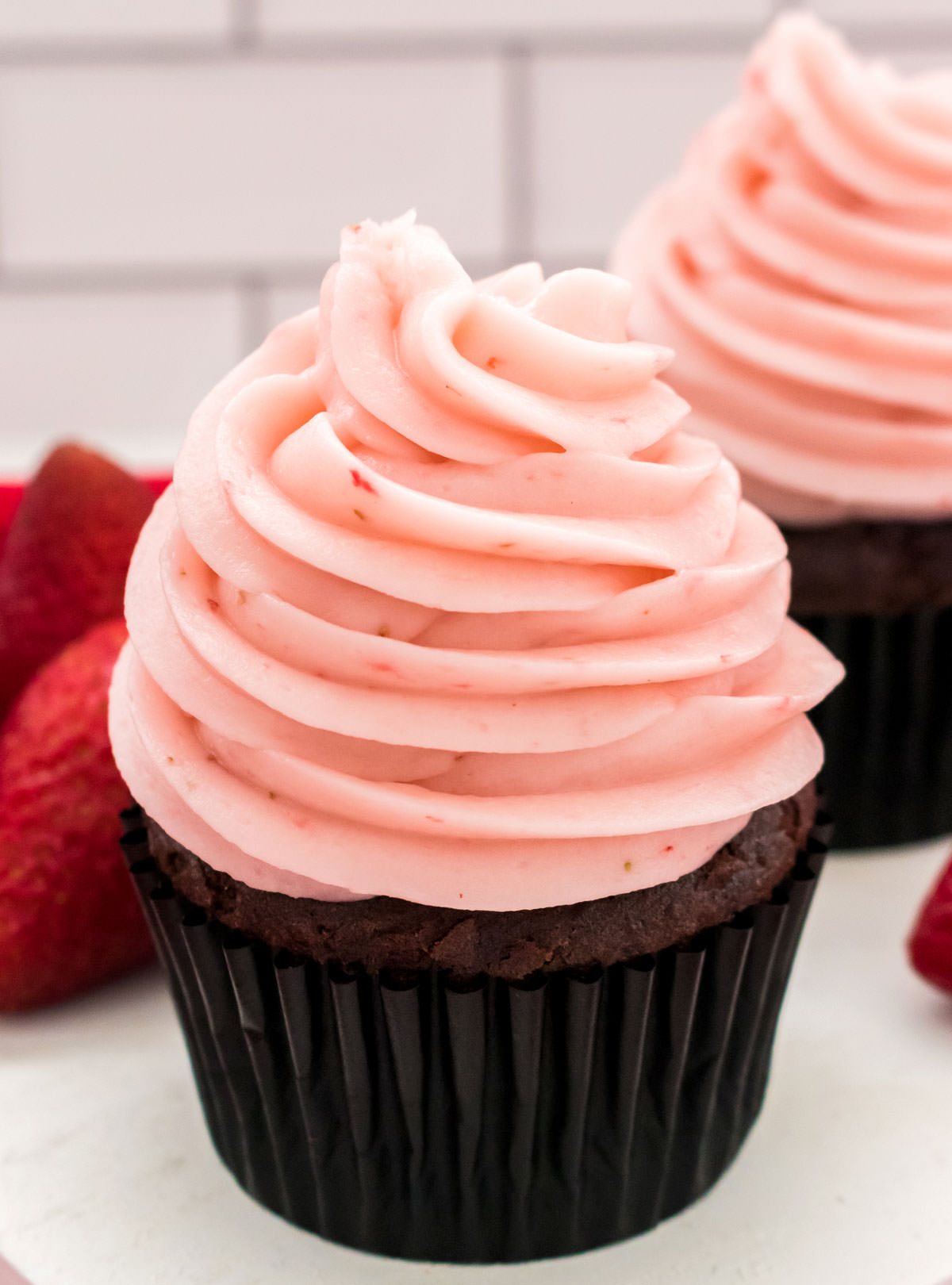 Closeup on two chocolate cupcakes topped with The Best Strawberry Buttercream Frosting sitting next to a stack of fresh strawberries.