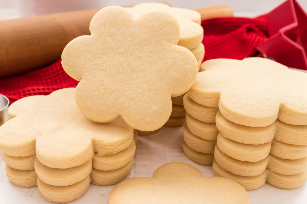 Closeup on three stacks of Sugar Cookies sitting on a white table in front of a red towel, a cookie cutter and a rolling pin.