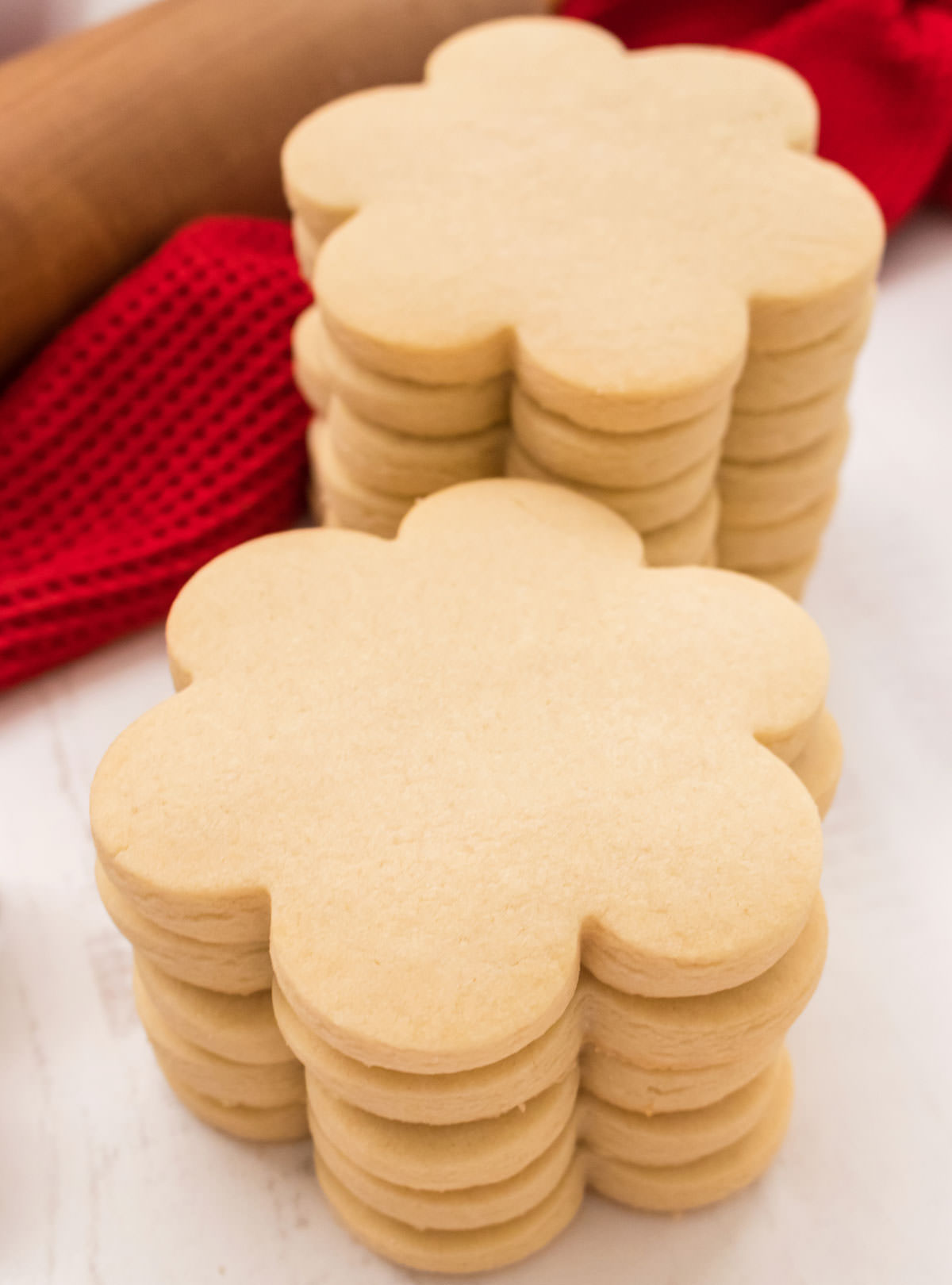 Closeup on two stacks of perfect flower sugar cookies made from our The Best Sugar Cookie Recipe sitting in front of a red towel and a wooden rolling pin.