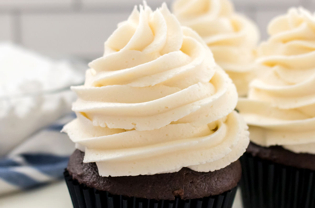 Closeup on three cupcakes topped with The Best Whipped Vanilla Frosting sitting on white table next to a glass bowl filled with powdered sugar.