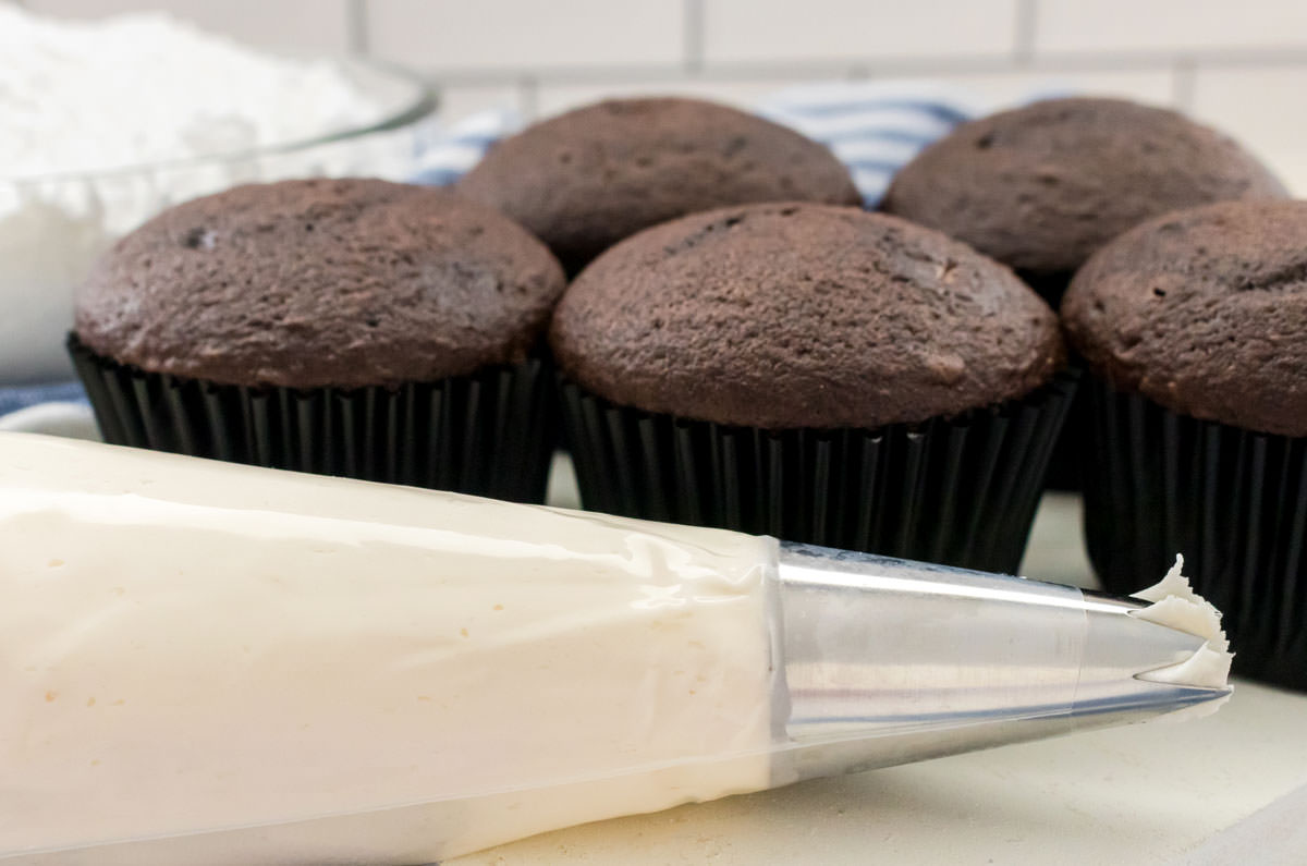 Closeup on a decorating bag filled with Whipped Vanilla Buttercream Frosting sitting in front of a batch of unfrosted chocolate cupcakes.