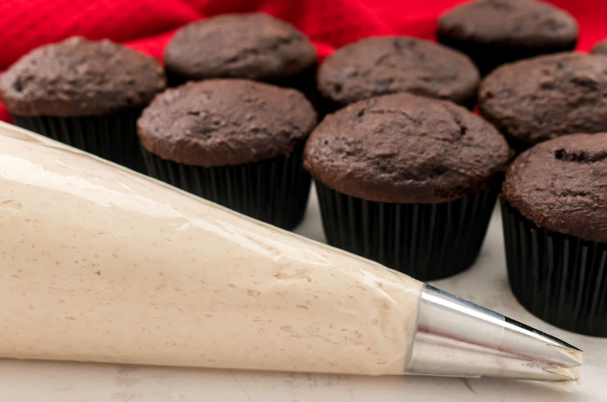 Closeup on a decorating bag filled with Brown Butter Frosting sitting in front of a batch of unfrosted chocolate cupcakes.