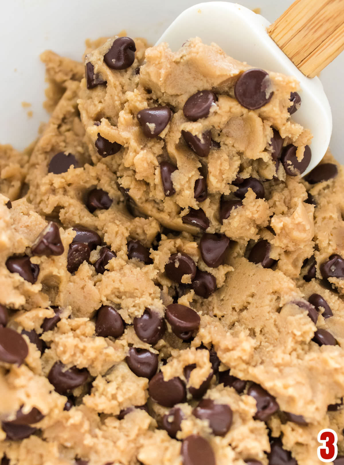 Closeup on a white mixing bowl filled with Brown Butter Chocolate Chip Cookie dough and a wooden spatula.