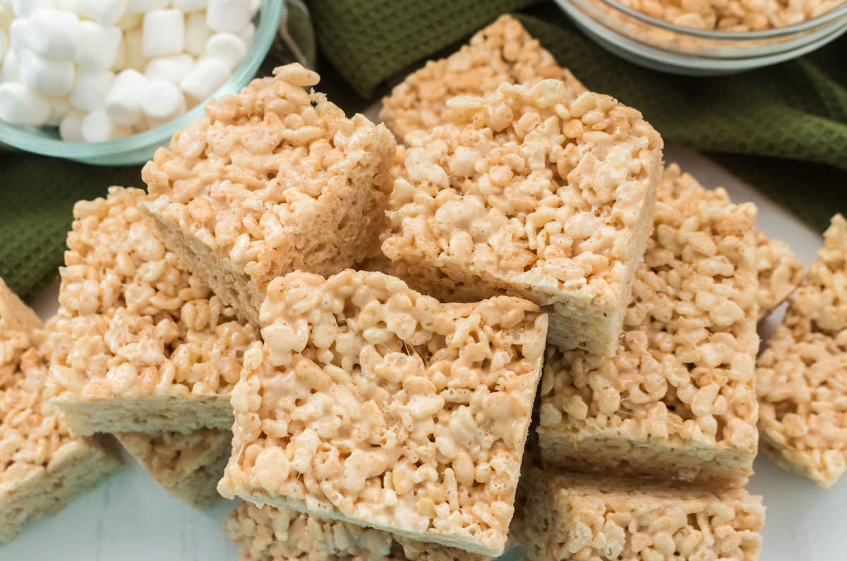 Overhead shot of a stack of Brown Butter rice Krispie Treats laying on a white surface with mini marshmallows and Rice Krispie Cereal in the background.