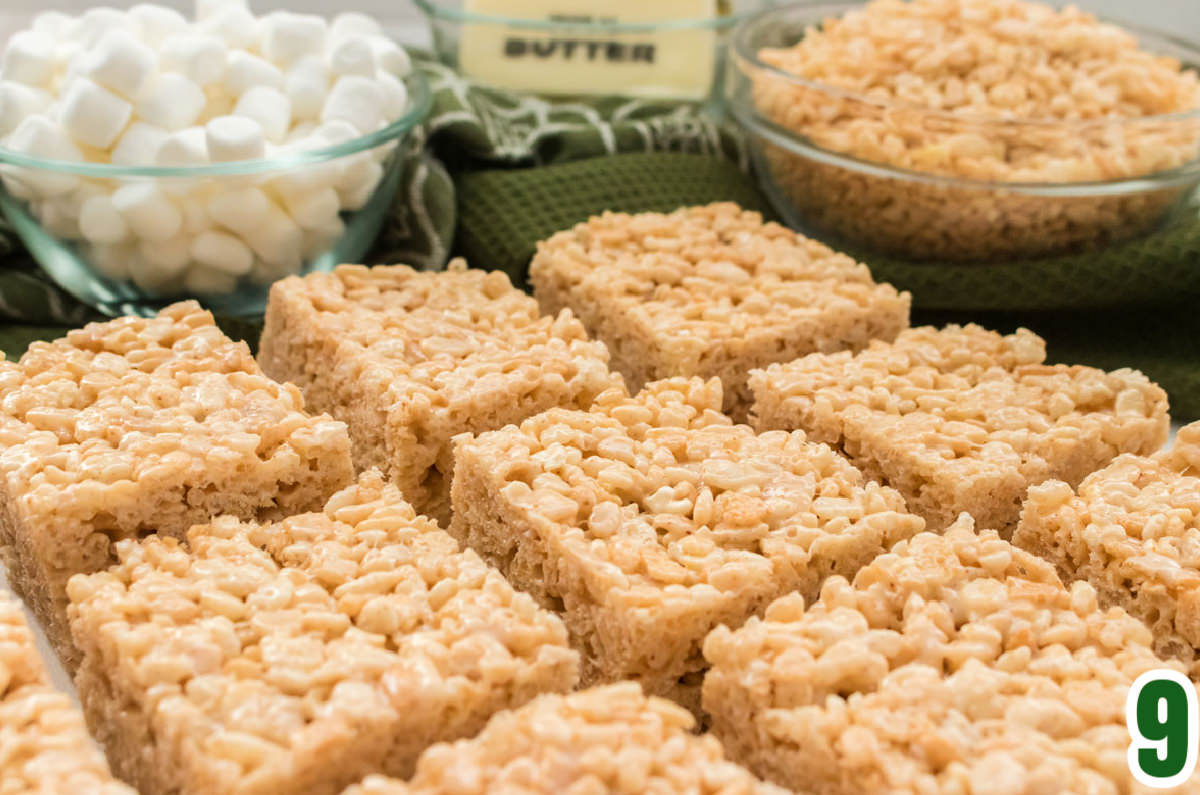 Closeup on nine Brown Butter Rice Krispie Treats laying in rows on a table with bowls of marshmallows and Rice Krispie Cereal in the background.