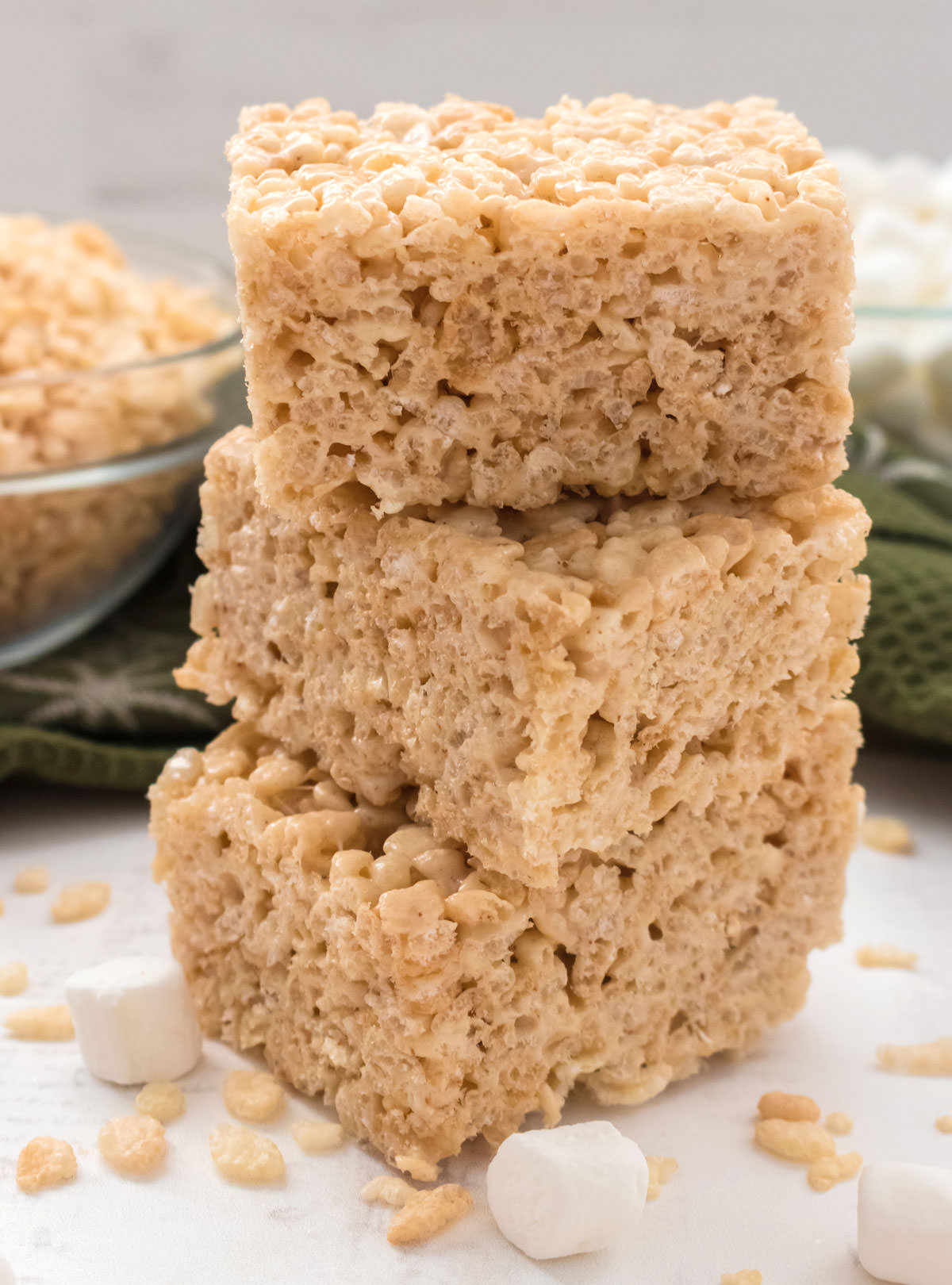 Closeup on a stack of Brown Butter Rice Krispie Treats sitting on a white surface in front of glass bowls filled with Rice Krispie Cereal and Mini Marshmallows.