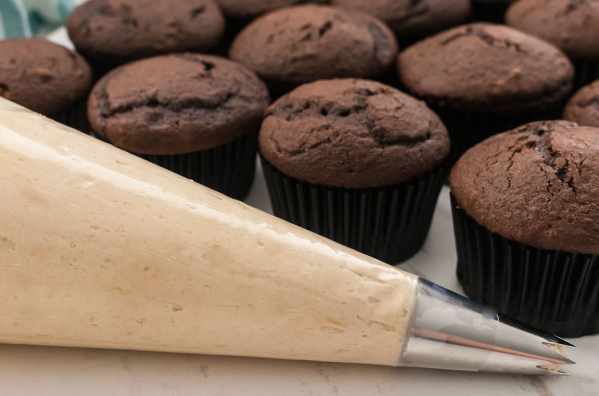 Closeup on a decorating bag filled with Brown Sugar Frosting sitting in front of a batch of unfrosted chocolate cupcakes.