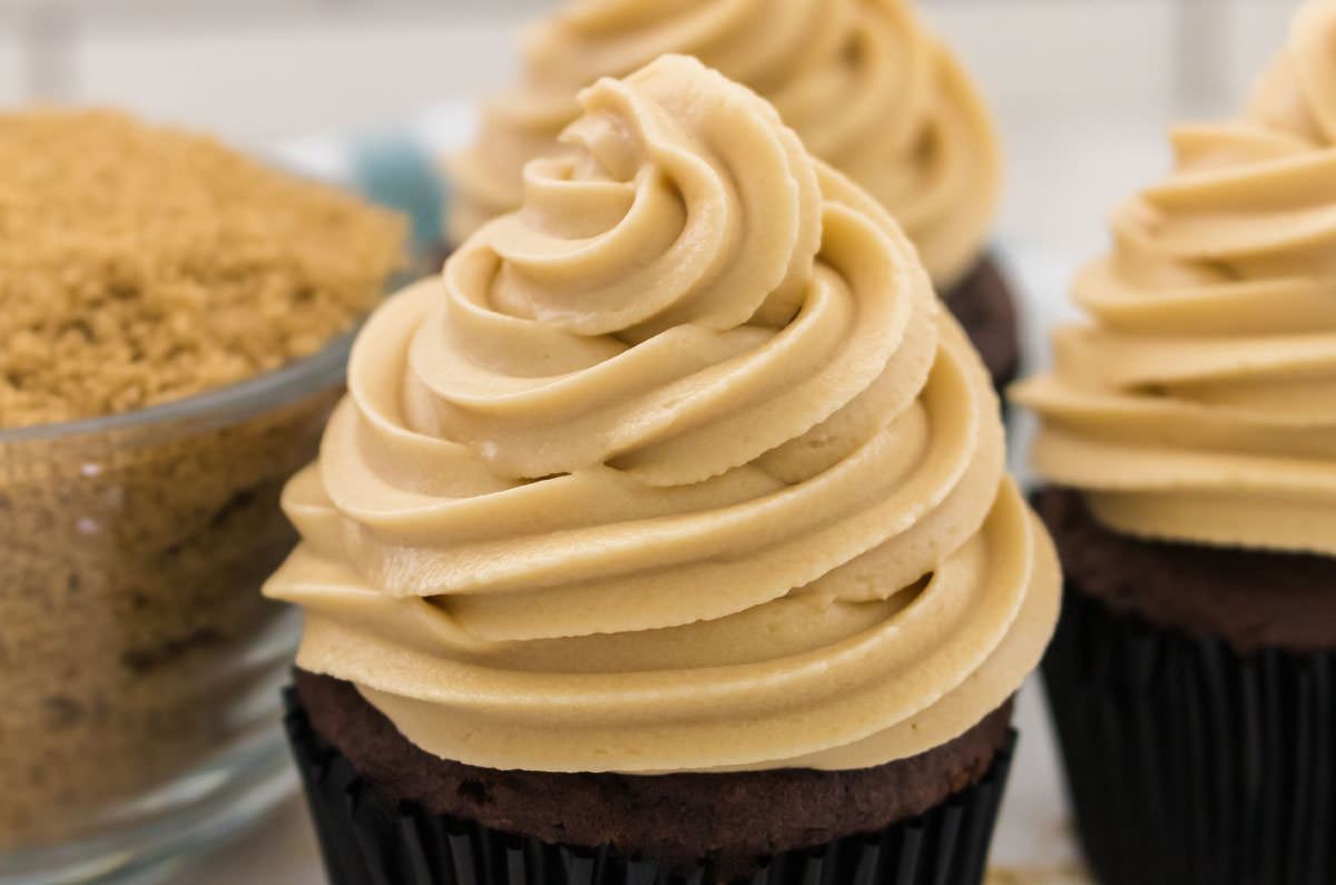 Closeup of three cupcakes topped with The Best Brown Sugar Cream Cheese Frosting sitting next to a glass bowl filled with Brown Sugar.