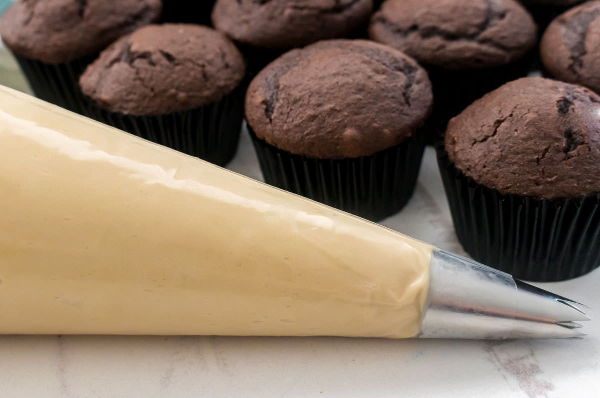 Closeup on a decorating bag filled with Brown Sugar Cream Cheese Frosting sitting in front of a batch of unfrosted Chocolate Cupcakes.