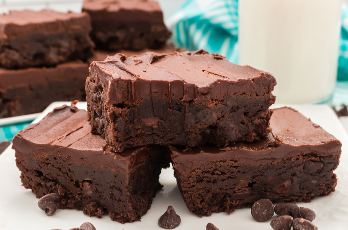 Brownies frosted with chocolate frosting sitting on a white plate in front of more brownies and a big glass of milk.