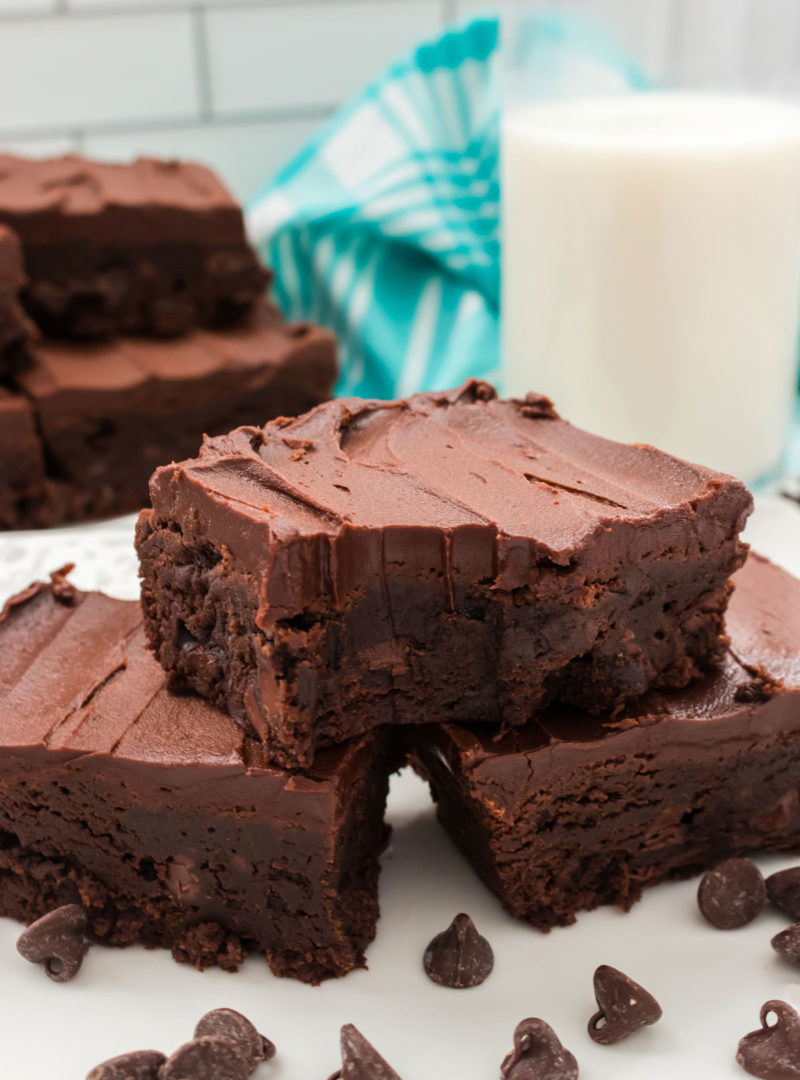 Three frosted brownies sitting on a white plate surrounded by chocolate chips and a tall glass of milk.