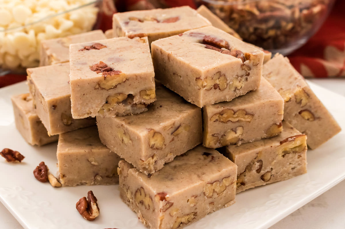 Closeup on a stack of Butter Pecan Fudge sitting on a white plate in front of glass bowls filled with pecans and White Chocolate Chips.