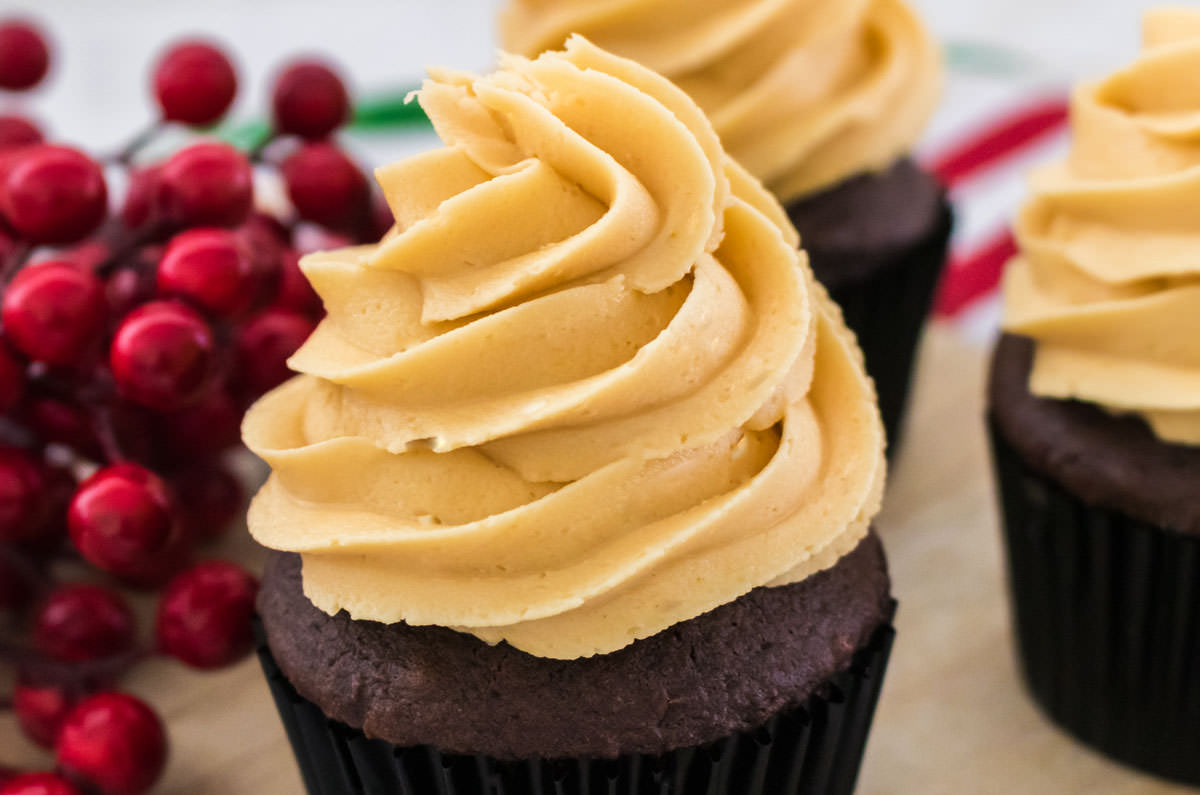 Closeup on three cupcakes topped with The Best Butter Rum Buttercream Frosting sitting on a cutting board next to a bunch of cranberries.