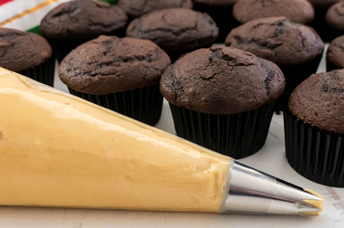 Closeup on a decorating bag filled with Butter Rum Frosting sitting in front of a batch of unfrosted chocolate cupcakes.