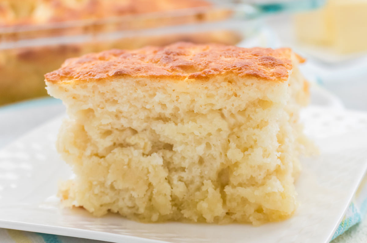 Closeup of a Butter Swim Biscuit sitting on a white plate on a white and blue table linen in front of a stick of butter and a pan of biscuits.